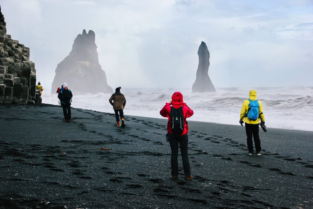 Gruppo di escursionisti in viaggio con Girolibero con k-way in una spiaggia nera camminano verso i faraglioni di Reynisdrangar, Islanda.